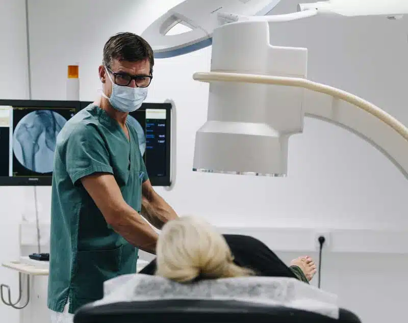 Bilde av Dr. Knut Egill Parrington Hansen som undersøker en kvinnelig pasient på et behandlingsrom. Pasienten ligger på en benk under en røntgen maskin. Legen har briller og munnbind på og holder hendene på pasienten - Stamcelleklinikken - Artrose