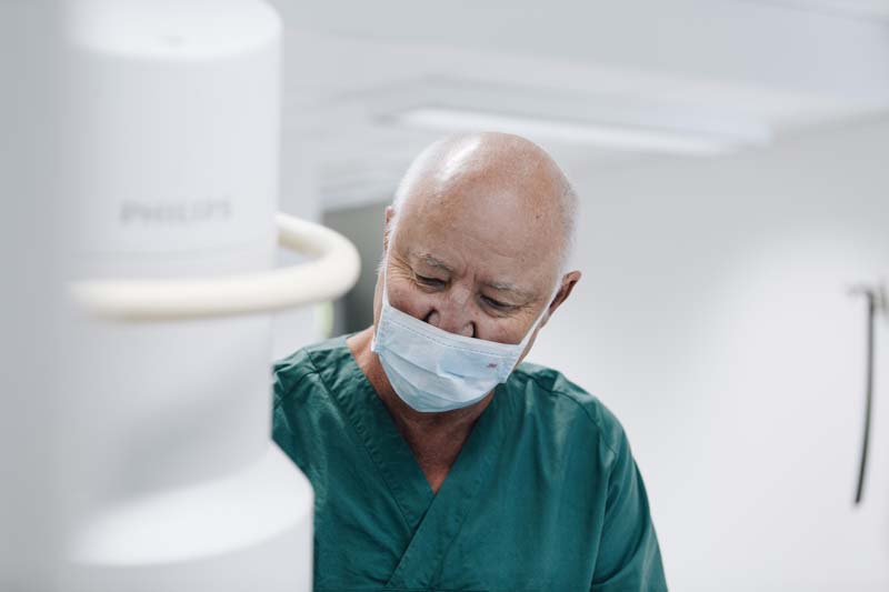 Bilde av Dr. Jørn Bremnes uten briller, med munnbind på. Han smiler ned mot pasienten på behandlingsrommet - Stamcelleklinikken - Artrose