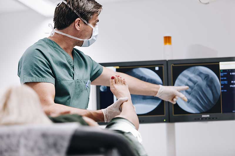 Bilde av Dr. Erik Bjørbæk bakfra som viser en kvinnelig pasient røntgenbilder på en monitor av pasientens fot, du ser knoklene til pasientens tær - Stamcelleklinikken - Artrose