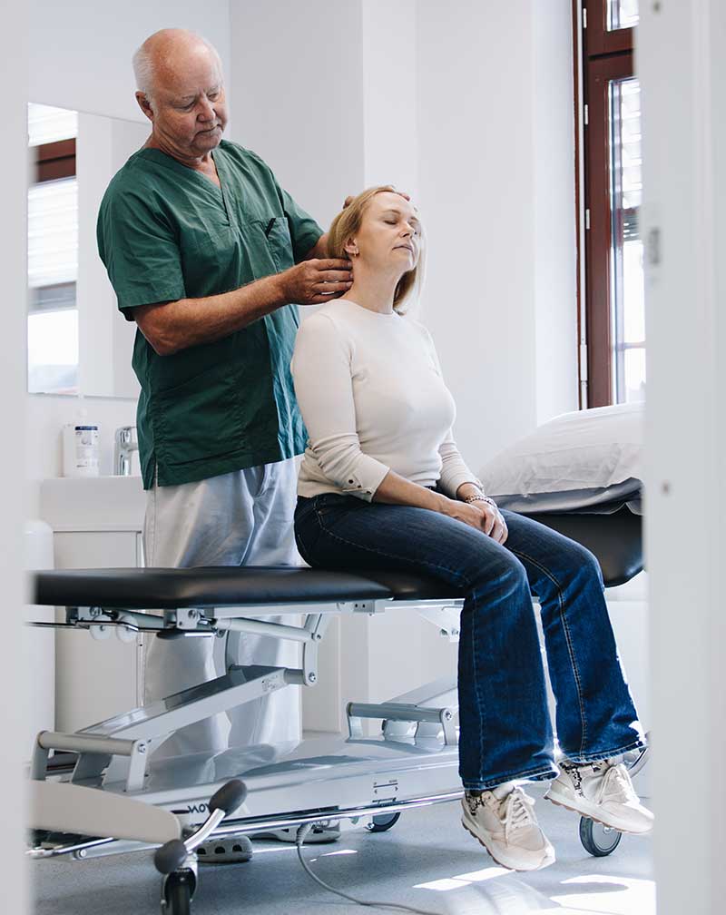 Bilde av Dr. Jørn Bremnes som undersøker en kvinnelig pasient. Pasienten sitter på en behandlingsbenk, legen undersøker muskulaturen i pasientens nakke - Stamcelleklinikken - Artrose